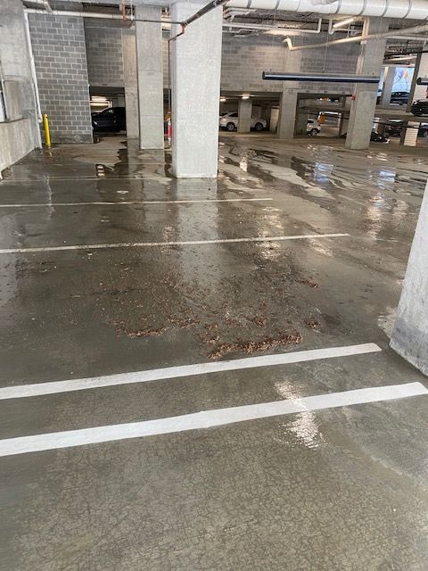 Wet concrete parking garage floor with water puddles and stains. White parking lines and support pillars are visible.