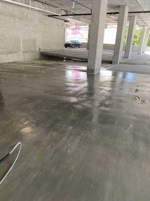 Interior of a concrete parking structure. Shiny gray floor reflects light, with pillars and a car visible in the background.