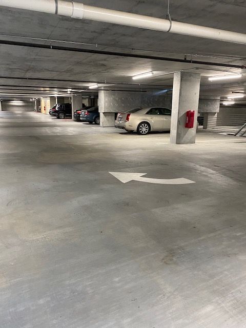 An empty, gray concrete parking garage with a white arrow on the floor. Several cars are parked in the background.