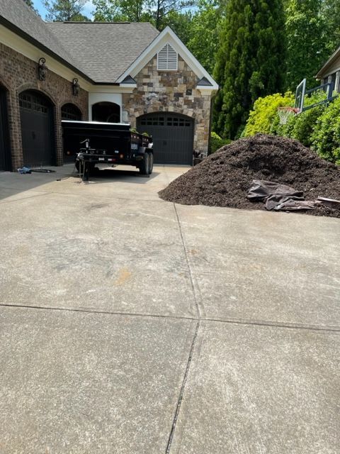 A driveway with a trailer, a large pile of mulch, and a garage. A house with stone and brick accents is in the background.