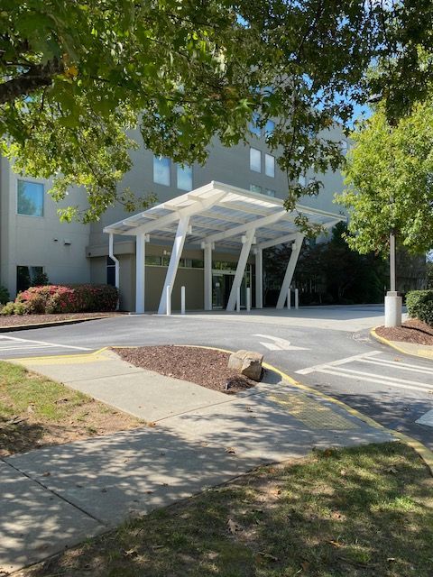 Building entrance with a white canopy. Asphalt driveway, trees, and shrubs.