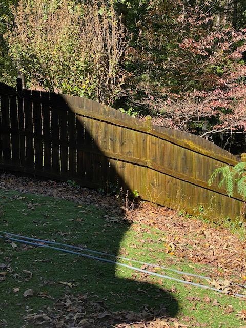 A weathered wooden fence along a grassy yard, partially shaded by sunlight, with trees in the background.