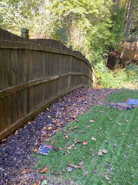 Wooden fence bordering a yard with green grass and a strip of black mulch covered in fallen leaves. Trees in the background.