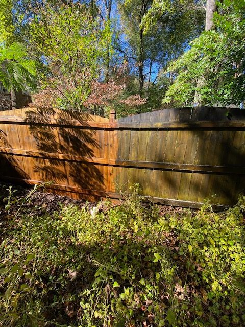 A wooden fence, half cleaned, contrasts between dark brown and green, surrounded by foliage and trees.