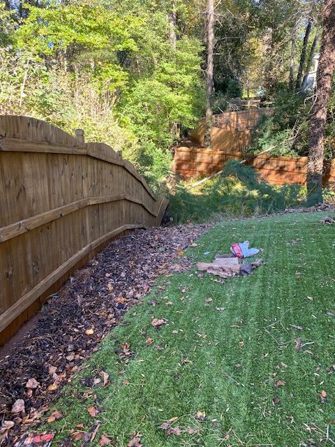 Wooden fence curving along a grassy area with mulch and leaves, leading into a wooded area with a higher fence.