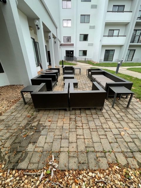 Patio furniture arranged on brick pavers outside a white building. Several dark gray sofas and tables are set up.