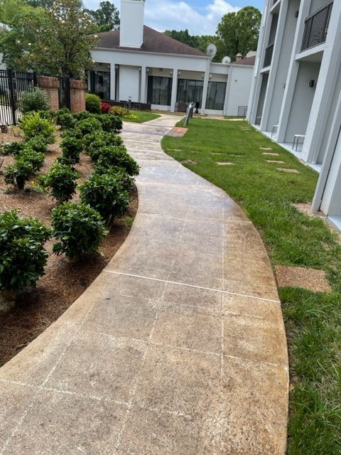 Concrete pathway curving through a landscaped area with shrubs, grass, and a building in the background. Overcast sky.