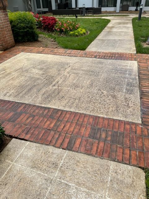 A brick-bordered concrete patio with a worn, textured surface adjoins a brick walkway, green grass, and flowers.