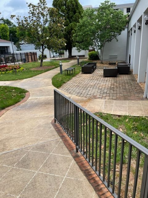 Exterior view of a paved courtyard with a metal railing. There's seating, a pathway, greenery, and a building in the background.