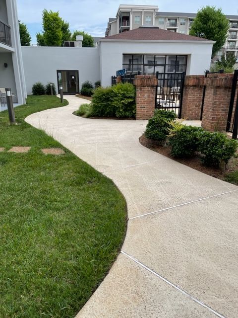 A winding concrete path leads to a building with a dark doorway and a gated area, bordered by green grass and shrubs.