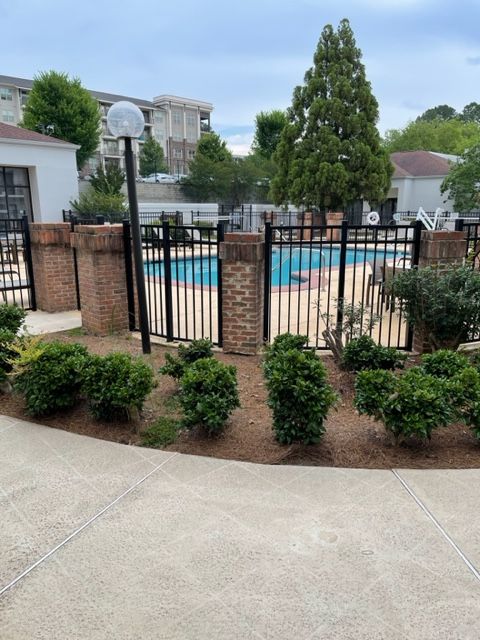 A fenced-in outdoor swimming pool surrounded by landscaping and brick pillars. Apartment buildings and trees are in the background.