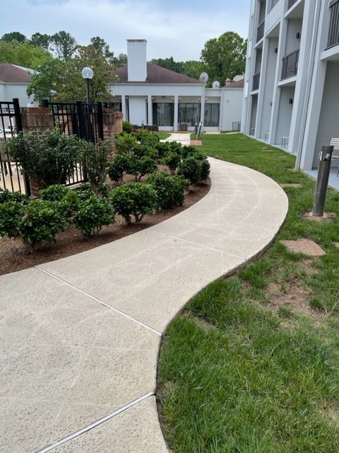 A curving concrete walkway bordered by green grass and shrubs leads toward a white building with a chimney.