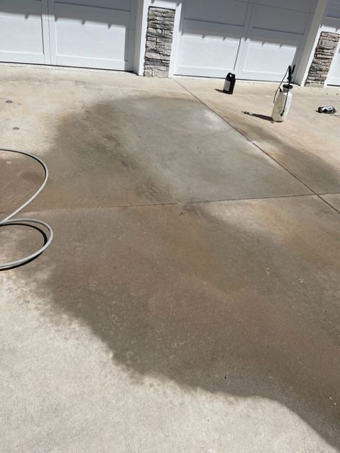Concrete driveway with dark stains, partially cleaned. A hose, sprayer, and cleaning supplies are visible near the garage doors.