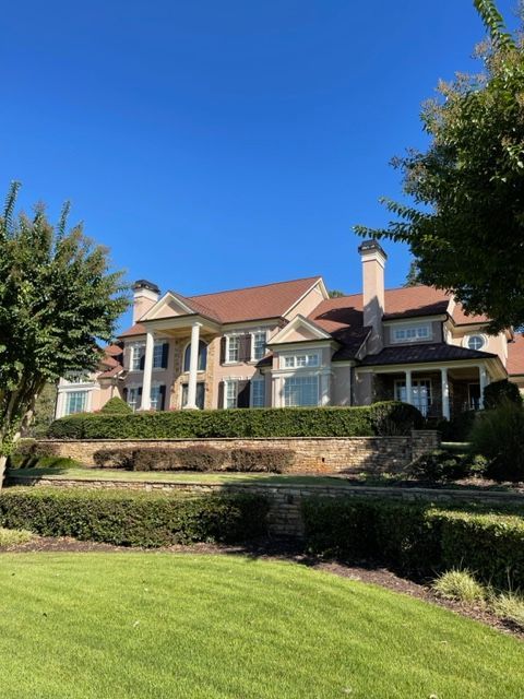 Large, beige house with a brown roof, columns, and manicured landscaping under a clear blue sky.