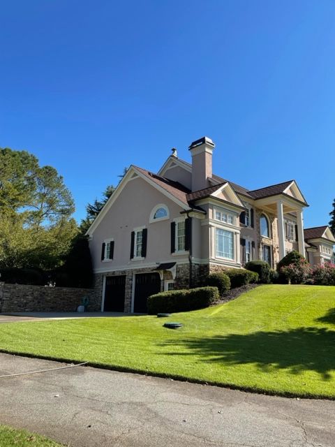 A large, two-story house with tan stucco walls, black shutters, and a brown roof, set on a grassy hill under a clear blue sky.