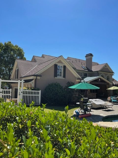 Backyard of a large house with a pool and patio furniture under a blue sky. Lush green bushes frame the foreground.