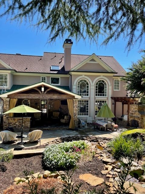 Rear view of a large house with a patio, outdoor kitchen, and landscaped yard with a pond. Green umbrellas, beige siding, and a blue sky.