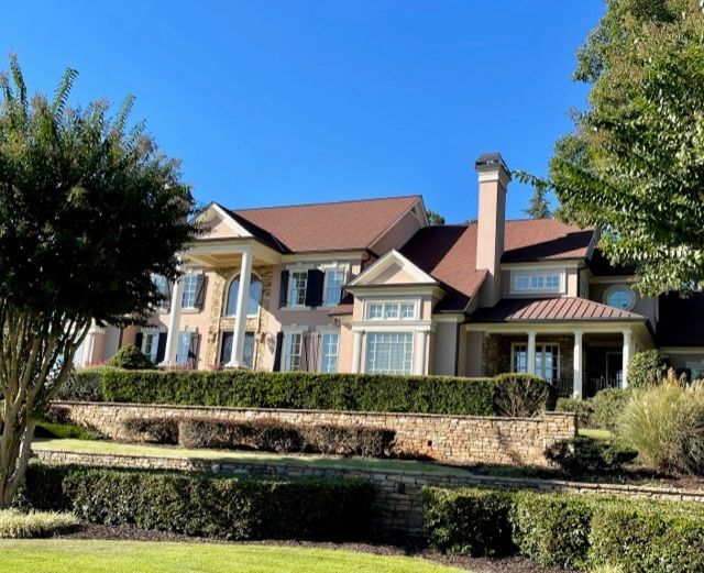 Luxurious two-story house with tan brick, brown roof, and columns. Green manicured hedges and lawns are in the foreground.