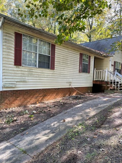 Beige house with red shutters and a concrete driveway. The house has a brick base and sits in a yard with trees.