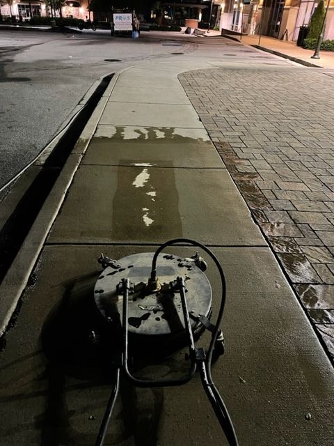 A sidewalk being cleaned with a pressure washer at night. The cleaned area is lighter than the dirty pavement.