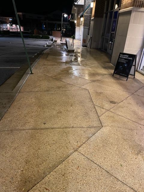 Wet sidewalk outside a building at night. An A-frame sign stands near the entrance.