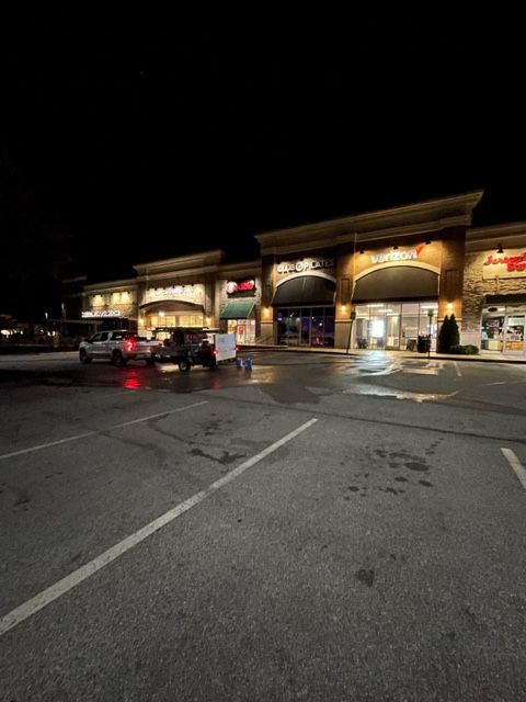 Night scene of a shopping plaza with storefronts lit up. Cars are parked in the empty lot in front.