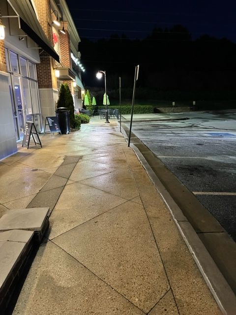 Exterior view of a wet sidewalk at night with a store and parking lot. Street lamps provide dim lighting.
