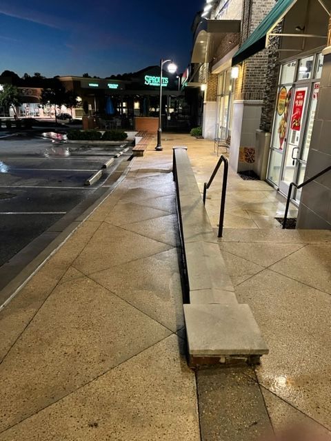 Exterior shot of a wet, paved sidewalk in front of a storefront at dusk. A low wall with a handrail extends along the sidewalk.