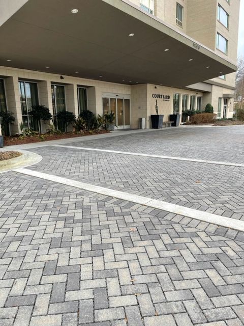Exterior view of a building with a herringbone brick driveway. A covered entrance leads to the building's glass doors.