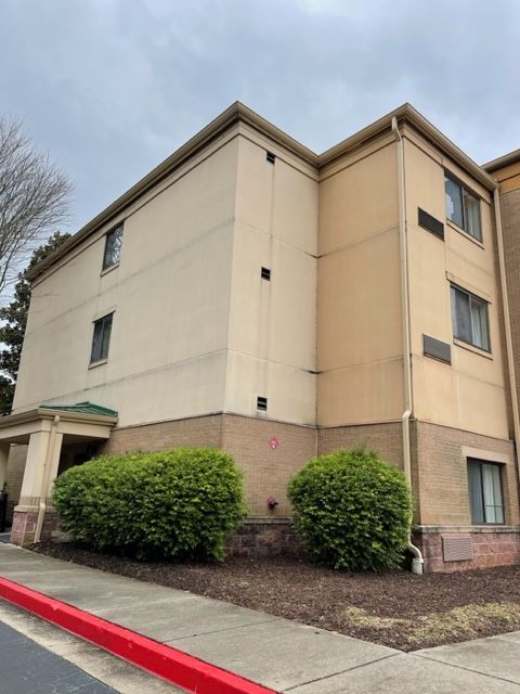 Three-story building with beige and brown exterior, bushes in front. Red curb and cloudy sky visible.
