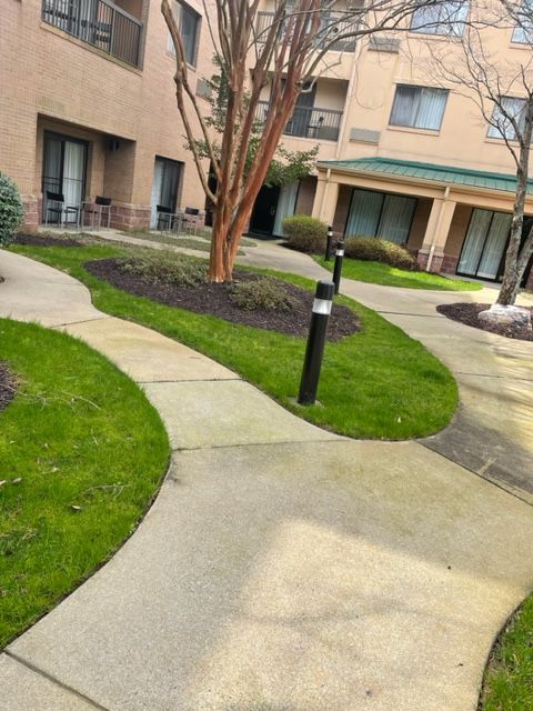 A concrete pathway winds through a courtyard with grass, trees, and a multi-story building. Several black light posts line the path.
