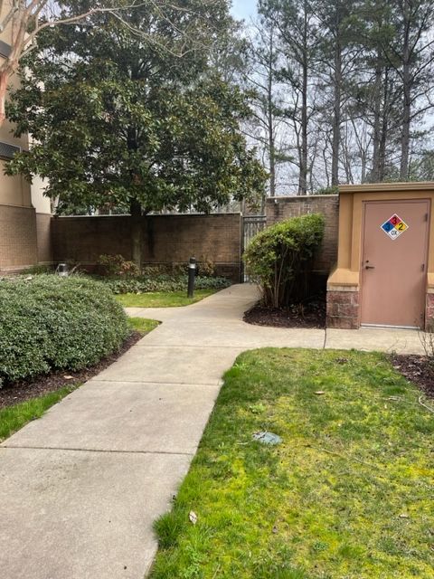 A concrete pathway curves through a green yard with a brick building and a brown door with hazard symbols.