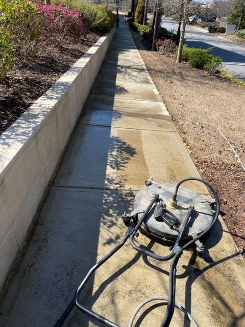 Sidewalk being power washed outdoors. Concrete shows a clean path next to a dirty portion; a pressure washer sits on the right.
