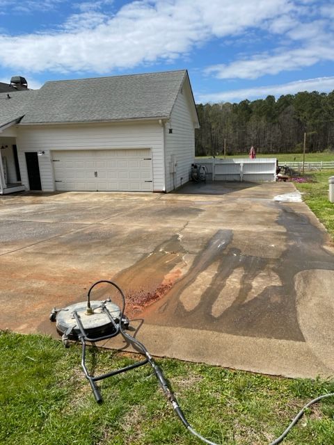 A concrete driveway being pressure washed in front of a white garage on a sunny day. A pressure washer is in the foreground.