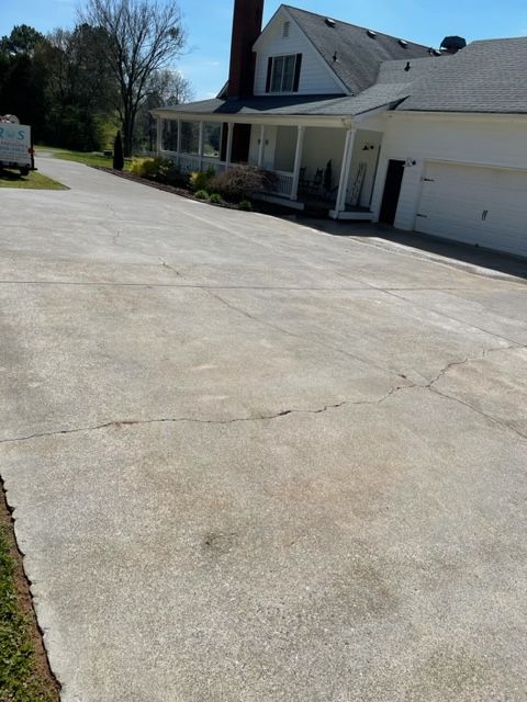 Concrete driveway leading to a white house with a porch and a garage. The driveway shows cracks and discoloration.