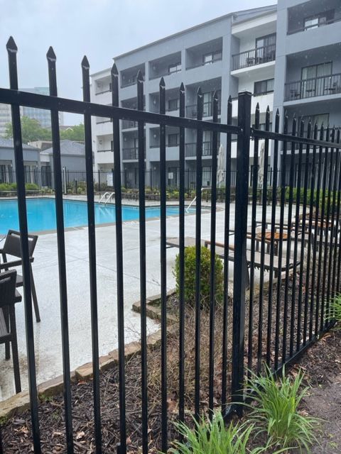 Black metal fence surrounding a blue swimming pool at an apartment complex. Building and chairs visible in the background.