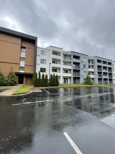 Modern apartment building with varied gray and brown facades under a cloudy sky; wet parking lot in foreground.