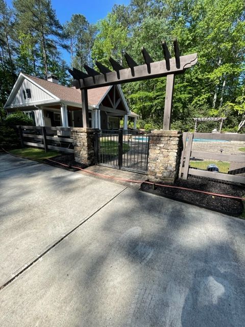 Stone pillars with a wooden pergola frame a black gate leading to a house and pool area.  The driveway is in the foreground.