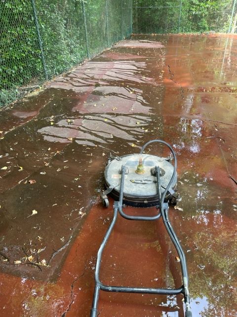 A person using a circular power washer on a red, wet tennis court covered in leaves, near a chain-link fence and trees.