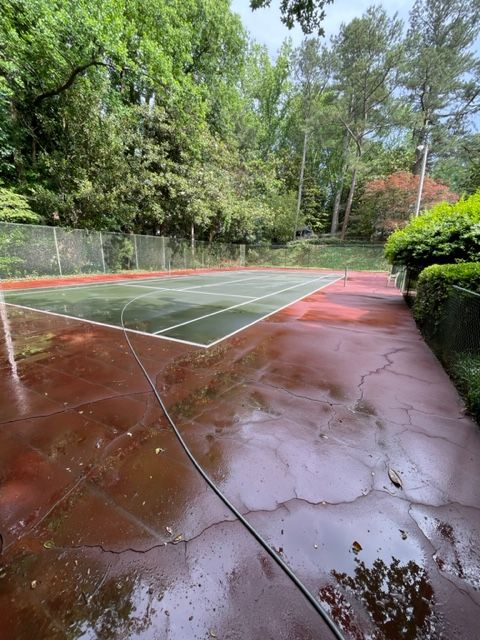 A wet tennis court, surrounded by green trees and a hedge, is being cleaned with a hose. The court's surface is red, with green sections.