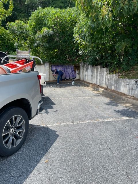A person working in a small parking area. A truck is parked nearby. The scene has a concrete wall and greenery.