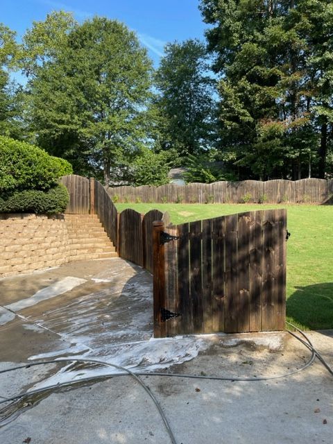 A wooden fence and gate in a backyard with grass, a concrete patio, and trees, showing the effects of cleaning with a pressure washer.