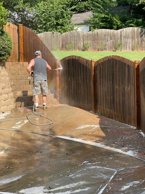 Man power washing a wooden fence in a backyard. The fence has arched sections; the ground is wet and covered in soap.