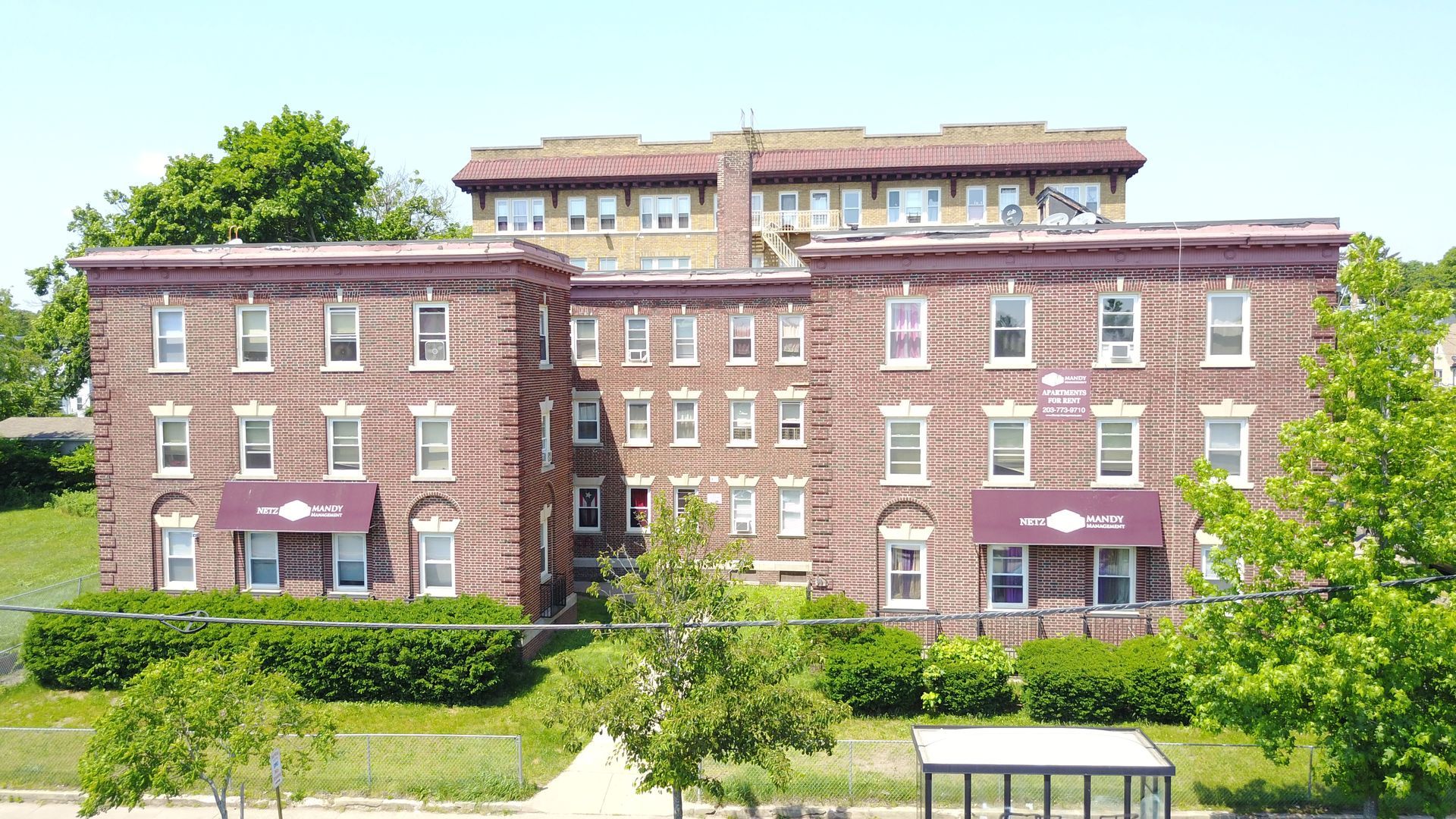 A multi-story red brick apartment building with symmetrical wings, awnings, and landscaped shrubs under a blue sky.