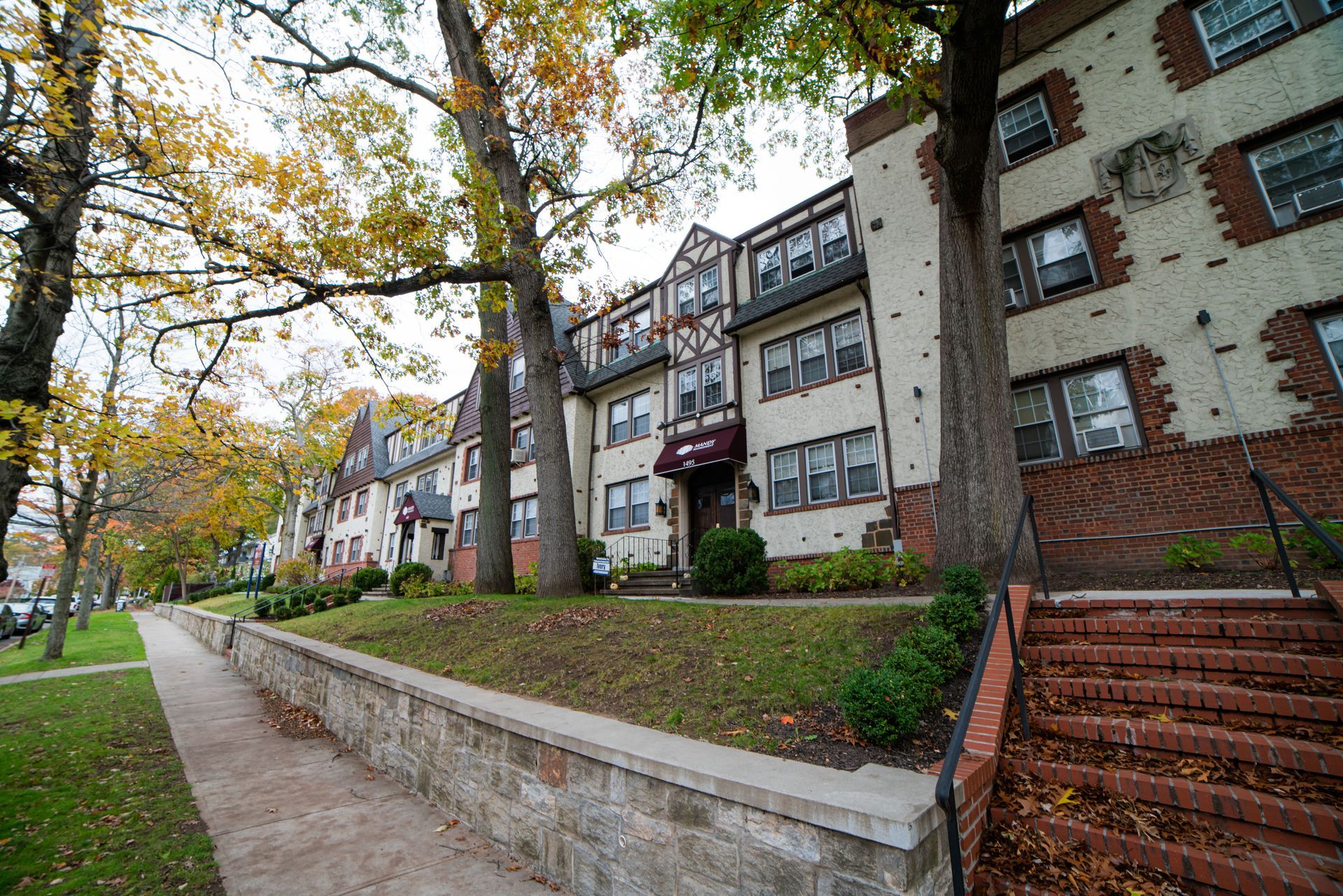 A multi-story brick apartment building with an exterior stone wall and steps on a tree-lined street during autumn.