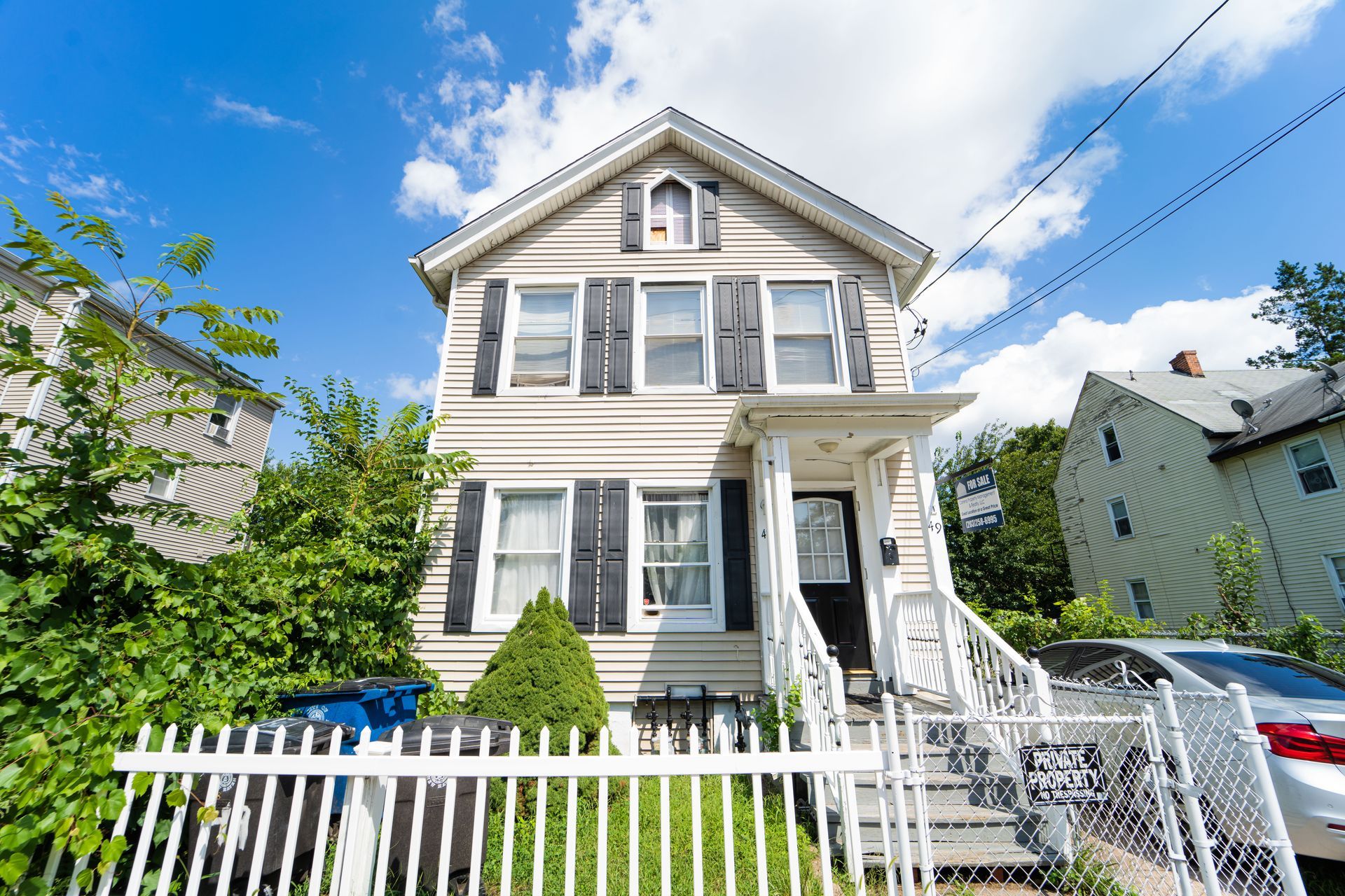 A two-story, beige-sided house with black shutters, a white porch, and a white picket fence under a partly cloudy sky.