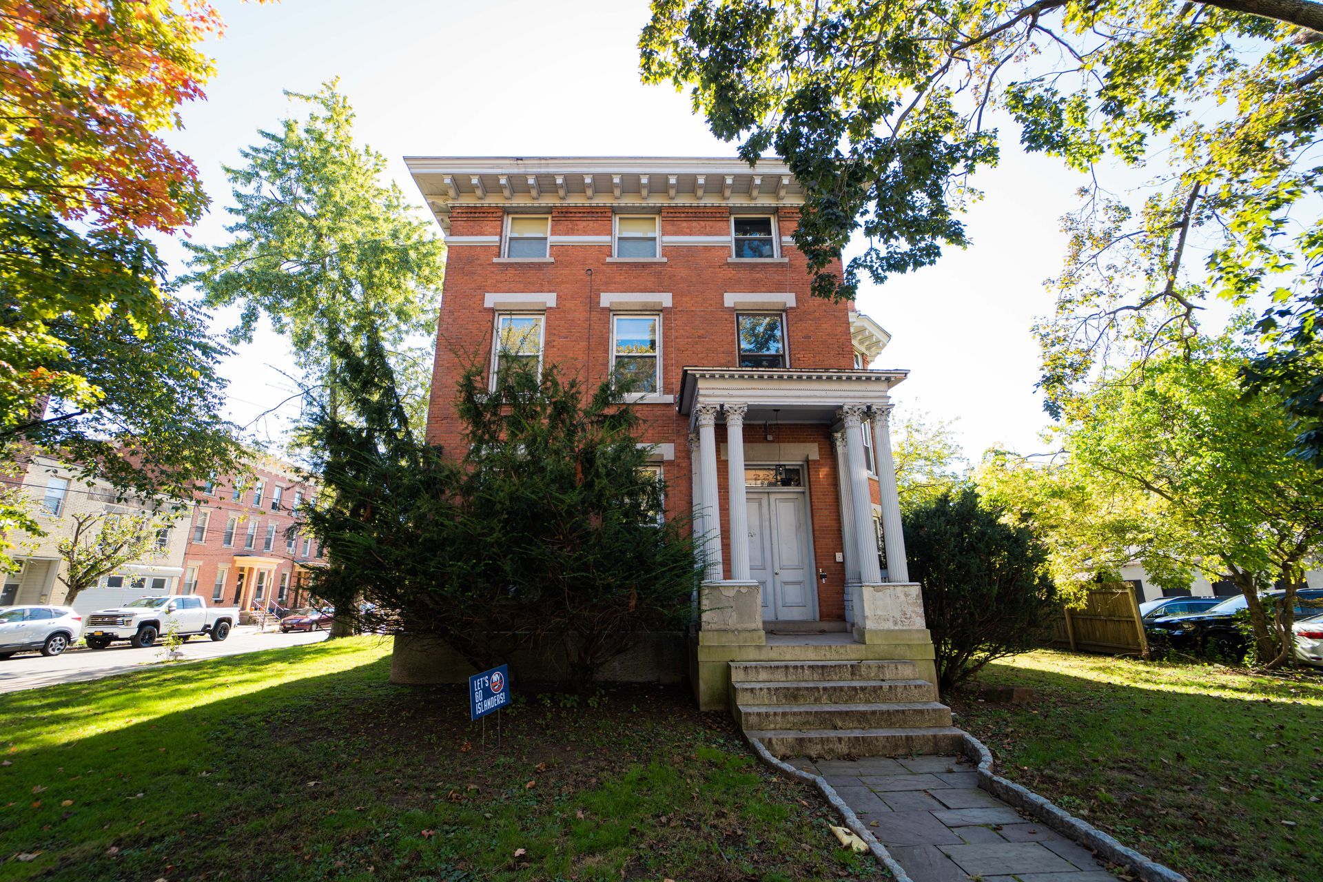 A three-story, red-brick house with white columns at the entrance, surrounded by trees on a sunny day.