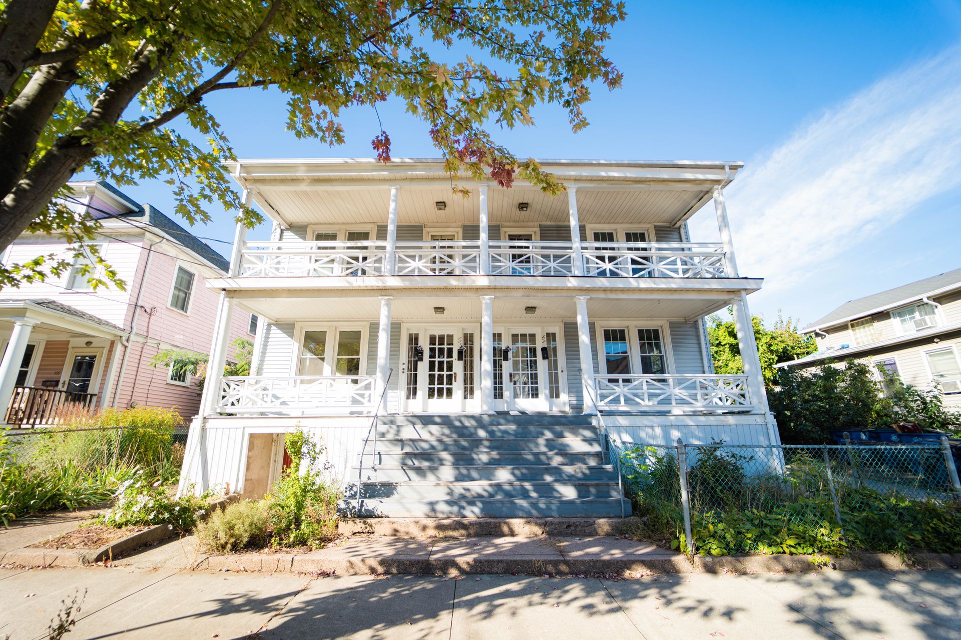 A two-story house with a white double-decker balcony, stairs leading to the entrance, and a tree branch in the foreground.