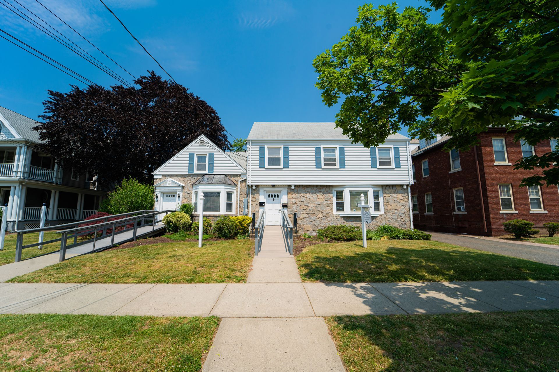 A two-story home with a stone and white siding exterior, featuring a center walkway and front lawn on a sunny day.
