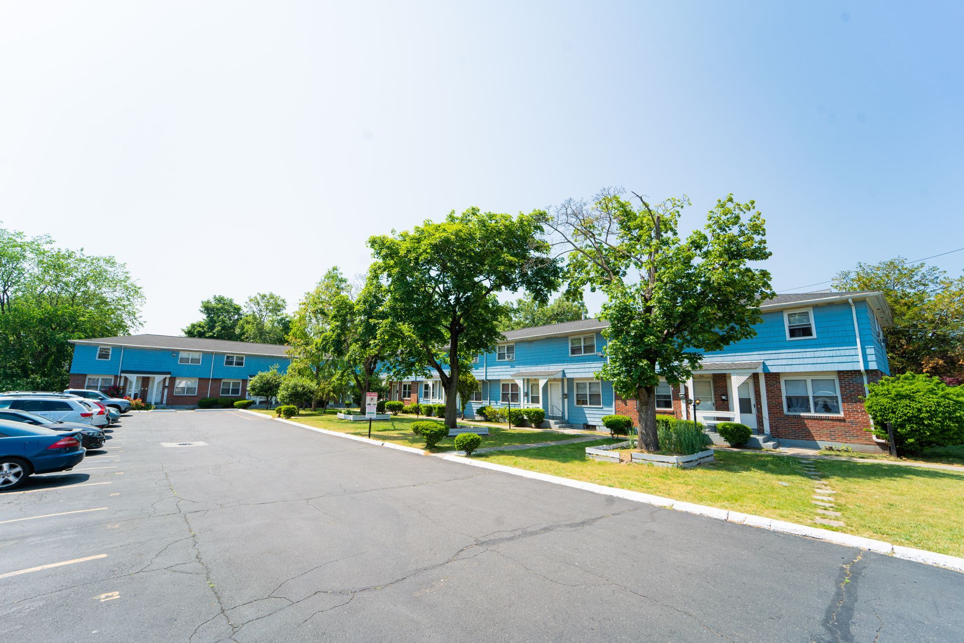 A parking lot in front of two-story, blue-sided apartment buildings with green trees on a sunny day.